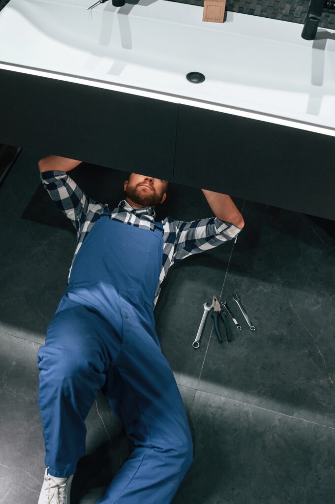 Tumbado en el suelo, arreglando el sifón. ​​Un fontanero de uniforme azul está trabajando en el baño